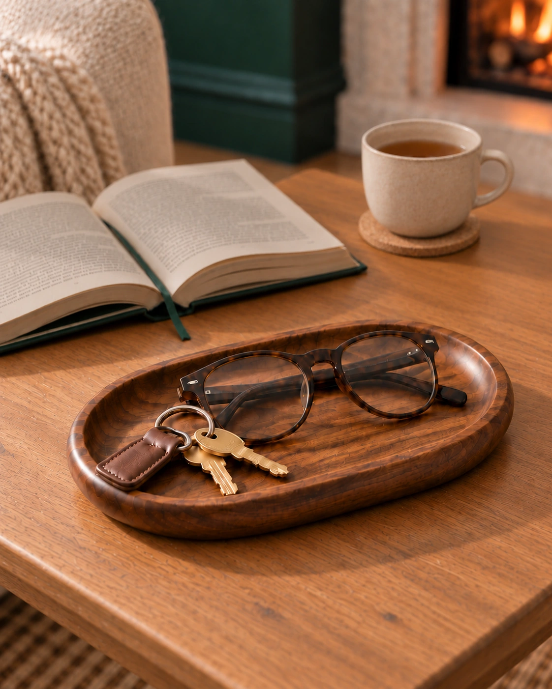 Oval wooden tray holding folded glasses and two keys on a wooden side table beside an open book and a cup in a winter interior.