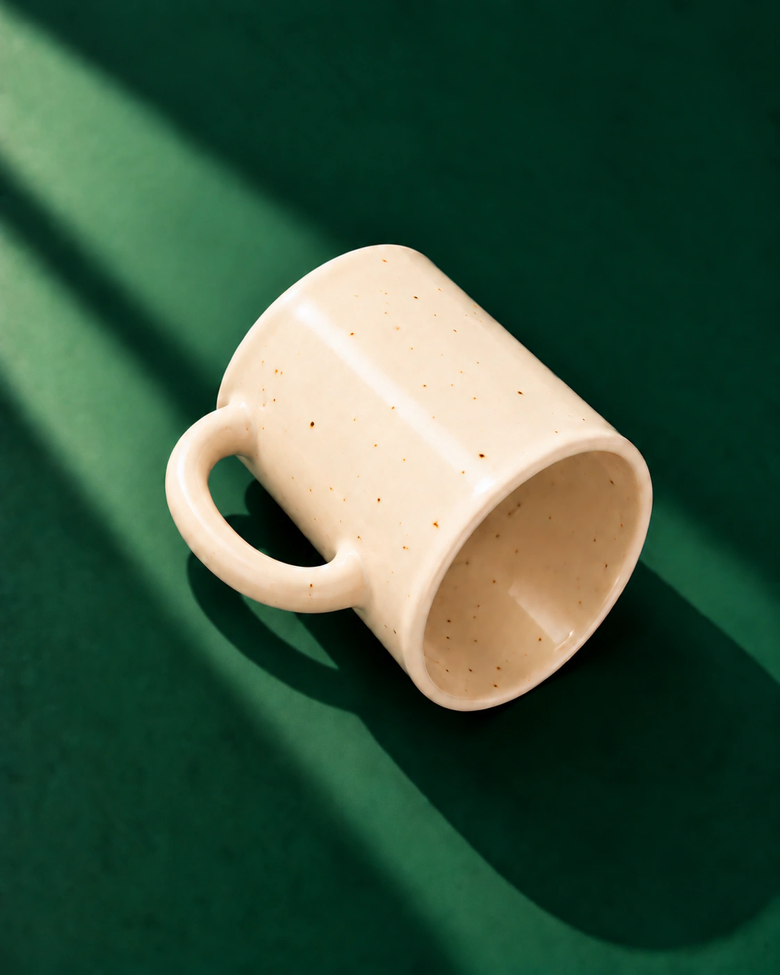 Ivory speckled mug resting diagonally on a deep green surface.