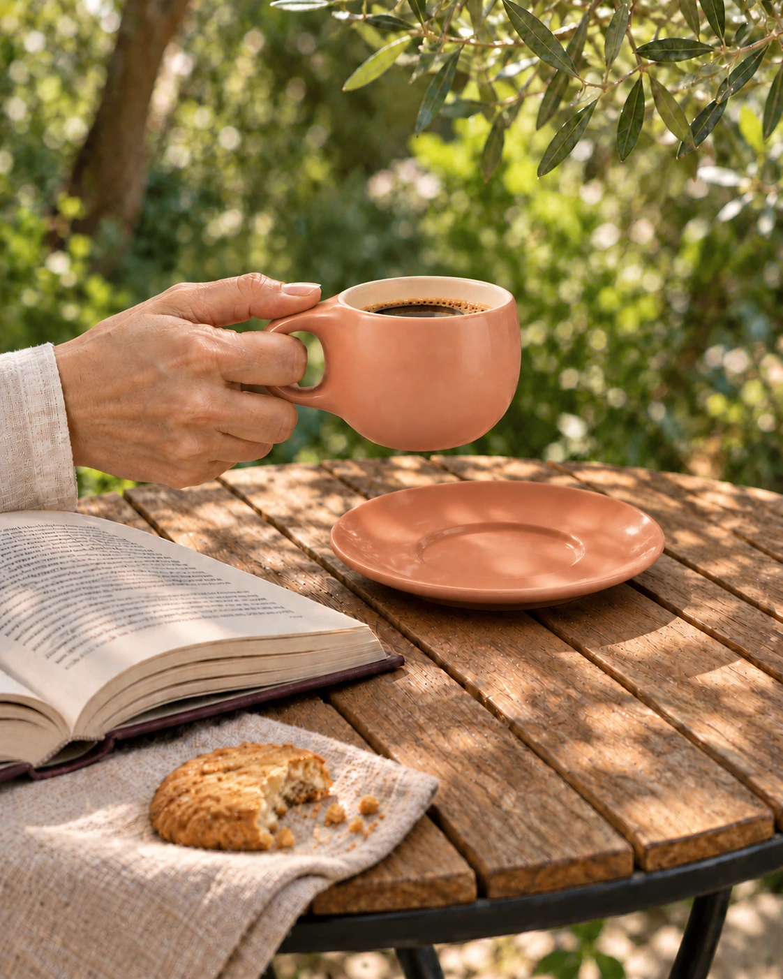 A peach ceramic cup held in one hand beside its saucer and an open book in a garden.